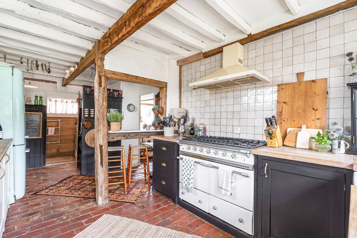 The kitchen: dark cabinetry, a white Lacanche, tiled splashback and whitewashed beams over a brick floor